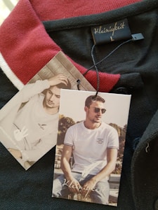 A clothing tag with the brand name 'Kleinigkeit' is attached to a garment featuring a red and black color scheme. The tags display images of a man, one sitting outdoors wearing sunglasses and a white t-shirt, while the other shows him indoors with a hand to his head.