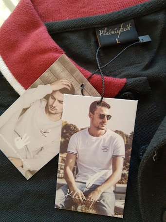 A clothing tag with the brand name 'Kleinigkeit' is attached to a garment featuring a red and black color scheme. The tags display images of a man, one sitting outdoors wearing sunglasses and a white t-shirt, while the other shows him indoors with a hand to his head.