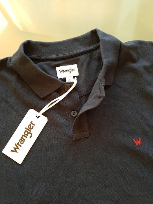 A dark-colored polo shirt with a collar and buttons, featuring a visible tag with the brand name 'Wrangler'. The red embroidered 'W' logo is on the chest area, and the tag is attached by a string.