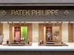 A luxury watch store display with the brand name Patek Philippe prominently featured at the top in gold letters. The window showcases an array of elegant watches on stands, surrounded by a soft, warmly lit interior. The backdrop includes artistic panels in shades of green and red, enhancing the refined appearance.