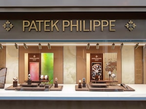 A luxury watch store display with the brand name Patek Philippe prominently featured at the top in gold letters. The window showcases an array of elegant watches on stands, surrounded by a soft, warmly lit interior. The backdrop includes artistic panels in shades of green and red, enhancing the refined appearance.
