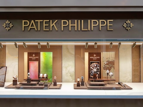 A luxury watch store display with the brand name Patek Philippe prominently featured at the top in gold letters. The window showcases an array of elegant watches on stands, surrounded by a soft, warmly lit interior. The backdrop includes artistic panels in shades of green and red, enhancing the refined appearance.