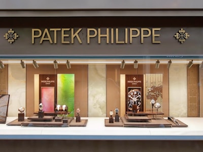 A luxury watch store display with the brand name Patek Philippe prominently featured at the top in gold letters. The window showcases an array of elegant watches on stands, surrounded by a soft, warmly lit interior. The backdrop includes artistic panels in shades of green and red, enhancing the refined appearance.