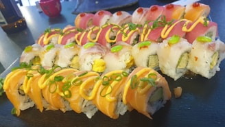 A platter of sushi rolls topped with various fresh toppings including salmon, tuna, and avocado, garnished with thinly sliced green onions and drizzled with a creamy sauce. The rolls are neatly arranged on a dark serving board, with vibrant colors showcasing the fresh ingredients.