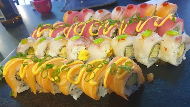 A platter of sushi rolls topped with various fresh toppings including salmon, tuna, and avocado, garnished with thinly sliced green onions and drizzled with a creamy sauce. The rolls are neatly arranged on a dark serving board, with vibrant colors showcasing the fresh ingredients.