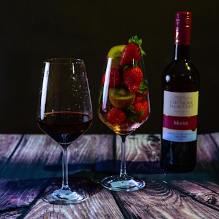 A wooden surface displays a bottle of red wine alongside two glasses. One glass contains red wine, while the other is filled with fresh strawberries and slices of kiwi. The scene is set against a dark, minimalist background, highlighting the vibrant colors of the fruit and the deep red of the wine.