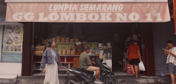 A small storefront with a sign above that reads 'Lunpia Semarang GG Lombok No 11'. The scene includes several people: a woman wearing a denim jacket and long skirt, a man sitting on a motorcycle, another person entering the shop with a bag, and a man checking a phone. The store displays stacks of packaged goods and posters advertising various food items.