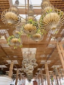 A ceiling installation features multiple large, ornate chandeliers composed of metal and adorned with green plants. The chandeliers hang from a wooden lattice structure that adds an intricate design element to the space. Natural light filters through the ceiling, creating an airy and open atmosphere.