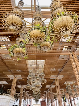 A ceiling installation features multiple large, ornate chandeliers composed of metal and adorned with green plants. The chandeliers hang from a wooden lattice structure that adds an intricate design element to the space. Natural light filters through the ceiling, creating an airy and open atmosphere.