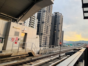 A railway station platform with train tracks leading into the distance. Tall residential buildings rise on the left against a backdrop of a softly lit sky during sunset. The architecture is modern, with clean lines and neutral colors.