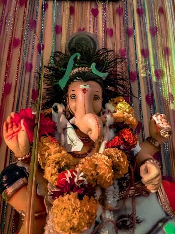 A vibrant statue of Lord Ganesha adorned with ornate garlands of marigold flowers. The statue features elaborate details, including a calm and serene expression and is placed against a decorative backdrop with heart patterns.