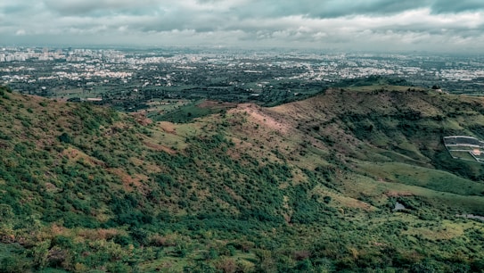 A lush green landscape with rolling hills covered in vegetation, overlooking a vast cityscape in the distance. The sky is overcast with thick clouds, casting a subdued light across the scene. The contrast between the natural landscape and the urban environment is striking.