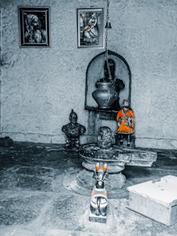 A small altar or shrine with religious artifacts and images. There are framed pictures on the wall depicting figures, a hanging metal vessel, and statues positioned on a stone platform. The central statue is a dark stone figure, possibly representing a deity, accompanied by a smaller colorful statue dressed in orange. The setting is rustic, with a stone floor and walls.