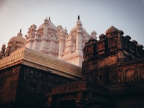 A majestic ancient temple with intricate carvings and domed structures rises into the sky. The architecture features ornate details with a blend of white and earth-toned stones. The lighting suggests early morning or late afternoon, casting soft shadows and a warm glow over the impressive facade.