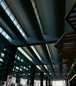 A modern architectural structure featuring a series of large, curved steel beams with skylight windows above. The space is expansive, with geometric lines and reflections of light on the glass surfaces of the walls.