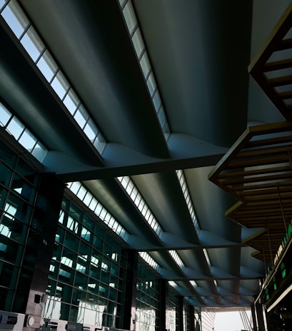 A modern architectural structure featuring a series of large, curved steel beams with skylight windows above. The space is expansive, with geometric lines and reflections of light on the glass surfaces of the walls.