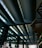 A modern architectural structure featuring a series of large, curved steel beams with skylight windows above. The space is expansive, with geometric lines and reflections of light on the glass surfaces of the walls.