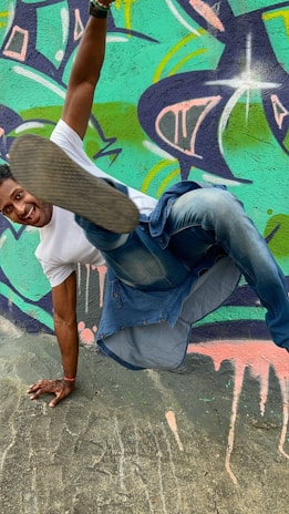 A person in a white t-shirt and jeans performs an impressive breakdance move against a colorful graffiti wall. The vibrant mural features abstract shapes and splashes of teal, purple, and pink.