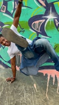 A person in a white t-shirt and jeans performs an impressive breakdance move against a colorful graffiti wall. The vibrant mural features abstract shapes and splashes of teal, purple, and pink.