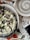 A pizza with a unique black crust topped with melted cheese, mushrooms, and herbs is placed on a white plate. The setting includes a round, empty bowl with a spiral design, a partial view of a smartphone displaying the time as 3:59, and a pair of keys on a terrazzo-style table.