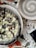 A pizza with a unique black crust topped with melted cheese, mushrooms, and herbs is placed on a white plate. The setting includes a round, empty bowl with a spiral design, a partial view of a smartphone displaying the time as 3:59, and a pair of keys on a terrazzo-style table.