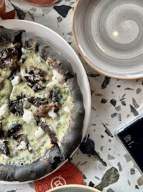 A pizza with a unique black crust topped with melted cheese, mushrooms, and herbs is placed on a white plate. The setting includes a round, empty bowl with a spiral design, a partial view of a smartphone displaying the time as 3:59, and a pair of keys on a terrazzo-style table.