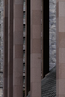 Numerous vertical concrete columns with a rectangular shape are aligned in parallel, creating a structured and symmetrical appearance. In the background, a wall of gray bricks is visible along with small, partially visible windows. There is also a slanted, grid-patterned glass structure at the bottom right corner, adding a geometric contrast to the scene.