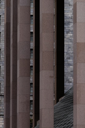 Numerous vertical concrete columns with a rectangular shape are aligned in parallel, creating a structured and symmetrical appearance. In the background, a wall of gray bricks is visible along with small, partially visible windows. There is also a slanted, grid-patterned glass structure at the bottom right corner, adding a geometric contrast to the scene.
