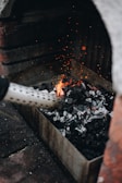 A tray filled with charcoal is being ignited with a fire starter, producing small flames and orange sparks. The setup appears to be in a grill or hearth environment, with bricks surrounding the area.
