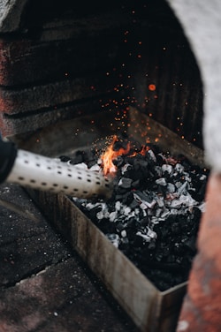 A tray filled with charcoal is being ignited with a fire starter, producing small flames and orange sparks. The setup appears to be in a grill or hearth environment, with bricks surrounding the area.