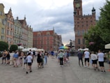 A busy European street scene featuring a group of people walking and gathering in a square. Colorful historic buildings with ornate architectural details and red-tiled roofs line the street. In the background, a large clock tower rises above the scene, beside which stands a statue. There are also several street vendors with umbrellas and outdoor seating areas with white canopies.