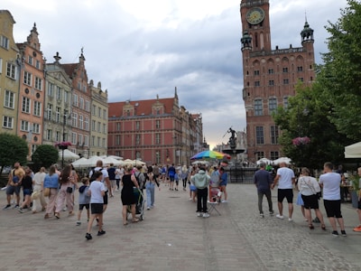 A busy European street scene featuring a group of people walking and gathering in a square. Colorful historic buildings with ornate architectural details and red-tiled roofs line the street. In the background, a large clock tower rises above the scene, beside which stands a statue. There are also several street vendors with umbrellas and outdoor seating areas with white canopies.