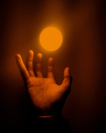 A hand with fingers slightly spread reaching upward towards a glowing, circular light source. The image is bathed in warm orange and brown tones, creating a dramatic and mysterious ambiance.