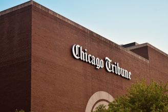 A large brick building featuring the name 'Chicago Tribune' in bold, white letters. The structure has a simple design with a few architectural details visible, such as arched patterns near the bottom. The sky is clear and blue, and some greenery is seen at the bottom right corner, suggesting nearby trees.