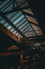 A dimly lit industrial interior featuring metal beams and a large skylight roof. Soft light filters through the glass panels, highlighting the structure's architectural elements.