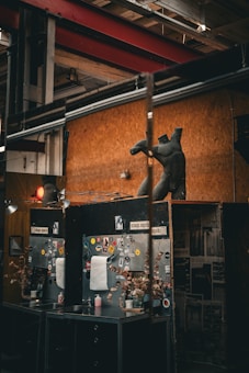 A room with industrial decor featuring a large dark sculpture resembling a torso without limbs. The walls are covered with wood paneling and posters. Two workstations are set up with mirrors, adorned with stickers and decorated with dried plants. Overhead lighting and exposed beams create a raw, artistic ambiance.