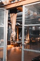 An entrance to a glass-walled room with metal frames. A beige curtain is tied back, and the sunlight is casting warm tones inside the room. There are several empty tables and chairs visible inside, with some candles on the tables, suggesting a calm and serene setting.