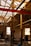 An industrial workshop space with a high ceiling supported by metal beams. Large windows allow natural light to illuminate the area. Equipment and tables are scattered throughout, with stacks of chairs and some electronic devices visible.