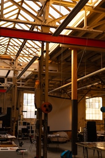 An industrial workshop space with a high ceiling supported by metal beams. Large windows allow natural light to illuminate the area. Equipment and tables are scattered throughout, with stacks of chairs and some electronic devices visible.