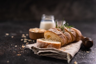 A rustic loaf of sliced bread is placed on a linen cloth, garnished with a sprig of rosemary. A jar of milk and a wooden bowl, possibly containing oats, are in the soft-focus background. Crumbly remnants are scattered across a dark, textured surface, creating a cozy and inviting atmosphere.