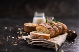 A rustic loaf of sliced bread is placed on a linen cloth, garnished with a sprig of rosemary. A jar of milk and a wooden bowl, possibly containing oats, are in the soft-focus background. Crumbly remnants are scattered across a dark, textured surface, creating a cozy and inviting atmosphere.