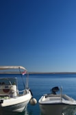Two motorboats are moored in calm, clear blue water under a clear sky. The boat on the left has a Croatian flag attached to the side, and both boats have visible registration numbers. The horizon is visible in the distance.