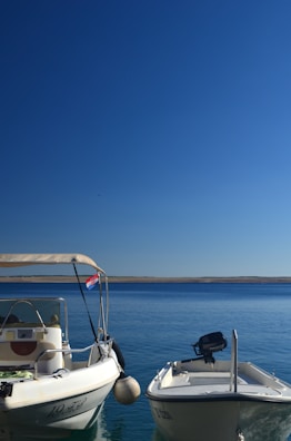 Two motorboats are moored in calm, clear blue water under a clear sky. The boat on the left has a Croatian flag attached to the side, and both boats have visible registration numbers. The horizon is visible in the distance.