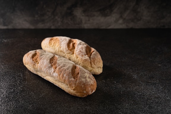Two loaves of rustic bread are placed side by side on a dark, textured surface. The bread has a golden-brown crust with distinct scoring marks, giving it a homemade and artisanal appearance.