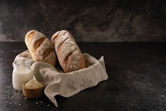 Two rustic loaves of bread rest on a linen cloth, accompanied by a small jar of milk and a wooden bowl of oats. The scene is set against a dark textured background, emphasizing a cozy and artisanal atmosphere.