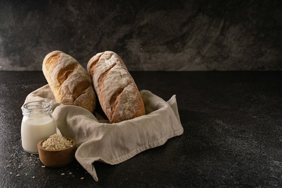 Two rustic loaves of bread rest on a linen cloth, accompanied by a small jar of milk and a wooden bowl of oats. The scene is set against a dark textured background, emphasizing a cozy and artisanal atmosphere.