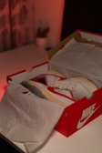 A pair of white sneakers with red accents placed inside a branded red shoebox on a white table. The sneakers are partially covered by white tissue paper, and there is a soft ambient red light in the background.