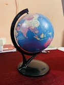 A globe displaying several countries including India, China, and surrounding regions is mounted on a black stand. It is positioned on a table with a dark red tablecloth. Some books and a pen are partially visible in the background, adding an academic or educational ambience.