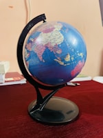 A globe displaying several countries including India, China, and surrounding regions is mounted on a black stand. It is positioned on a table with a dark red tablecloth. Some books and a pen are partially visible in the background, adding an academic or educational ambience.