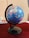 A globe displaying several countries including India, China, and surrounding regions is mounted on a black stand. It is positioned on a table with a dark red tablecloth. Some books and a pen are partially visible in the background, adding an academic or educational ambience.